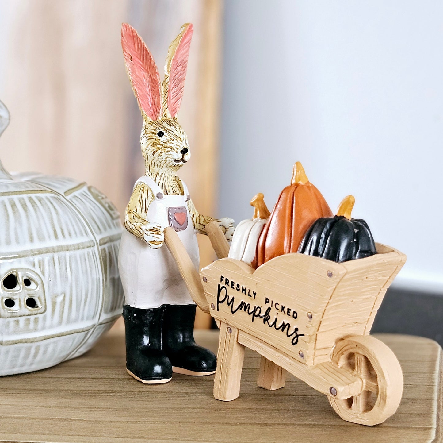 rabbit ornament dressed in dungerees and black boots holding a wheelbarrow with pumpkins and freshly picked pumpkins wording on the wheelbarrow