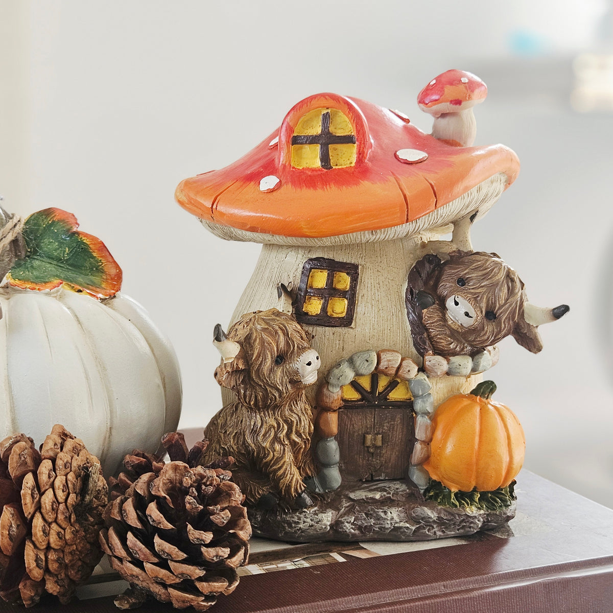 A decorative mushroom-shaped cottage figurine with a red-orange mushroom cap, cream base, small window, door, and two shaggy highland cow characters