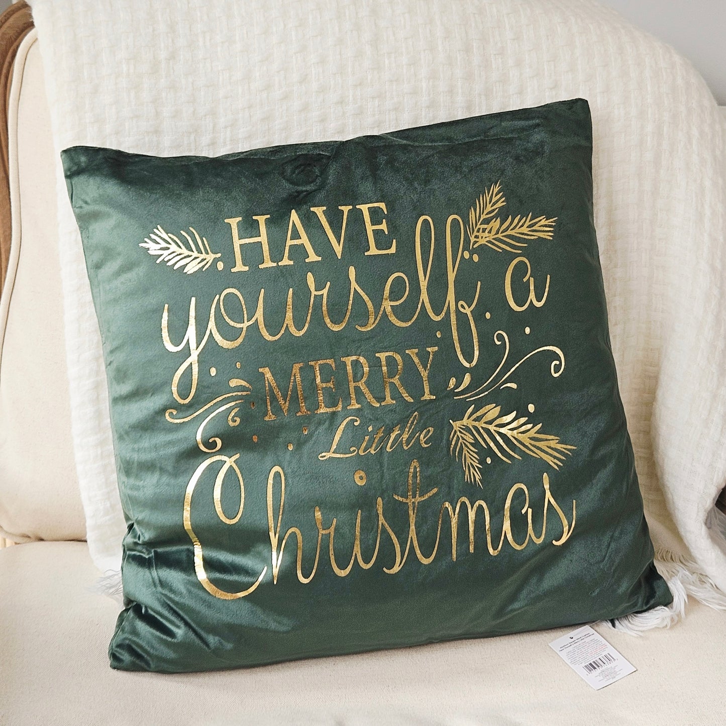 A square dark green velvet throw pillow with gold foil lettering that reads 'HAVE yourself a MERRY Little Christmas' and gold pine branch accents, resting on a cream-colored armchair.