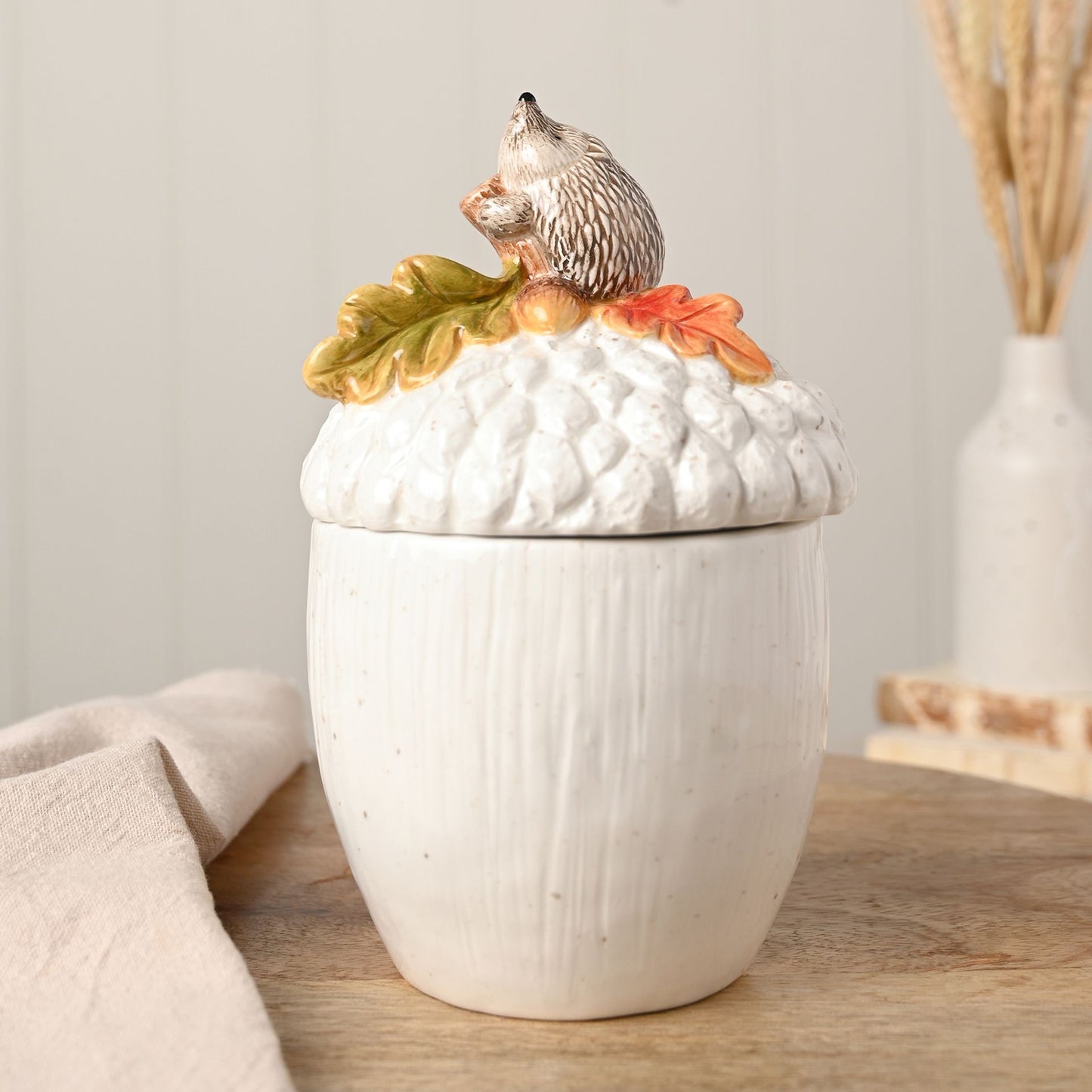 Acorn storage jar with hedgehog perched on the lid and colourful leaves 
