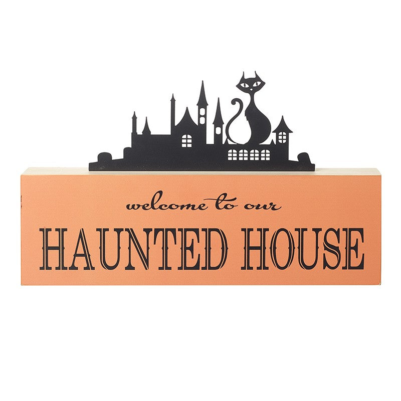 A rectangular orange decorative sign with black text reading 'welcome to our HAUNTED HOUSE' and a black silhouette skyline of a haunted house with a cat on top,