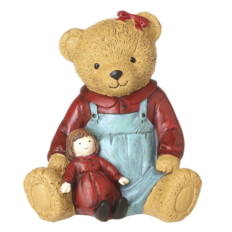 A teddy bear figurine wearing a red shirt and light blue overalls, sitting and holding a small doll with a red dress and brown hair,
