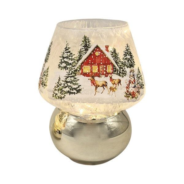 A festive table lamp with a clear, textured glass shade featuring a printed winter holiday scene of a red cabin, pine trees, and deer, set on a reflective silver base.
