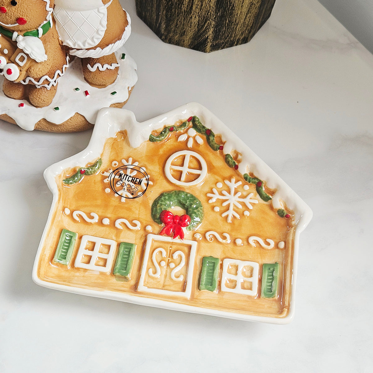 A ceramic gingerbread house-shaped dish with a warm tan base, white icing-style trim, green shutters and wreath accents, and a red bow