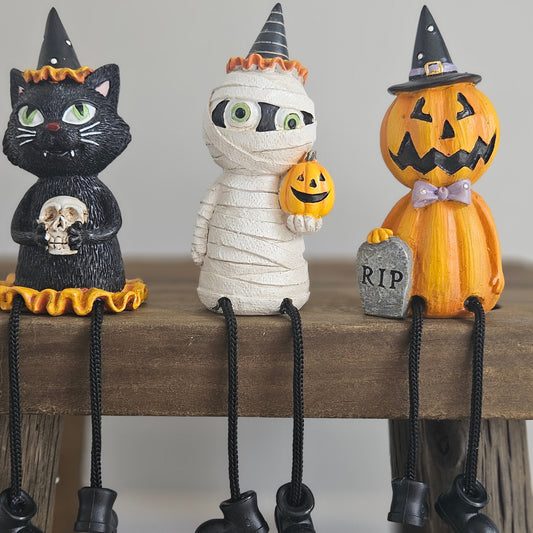 Three Halloween-themed decorative shelf sitter figurines a black cat holding a skull, a white mummy holding a small jack-o'-lantern, and an orange pumpkin character holding a tombstone. All have witch hats and dangling black boot-shaped feet 