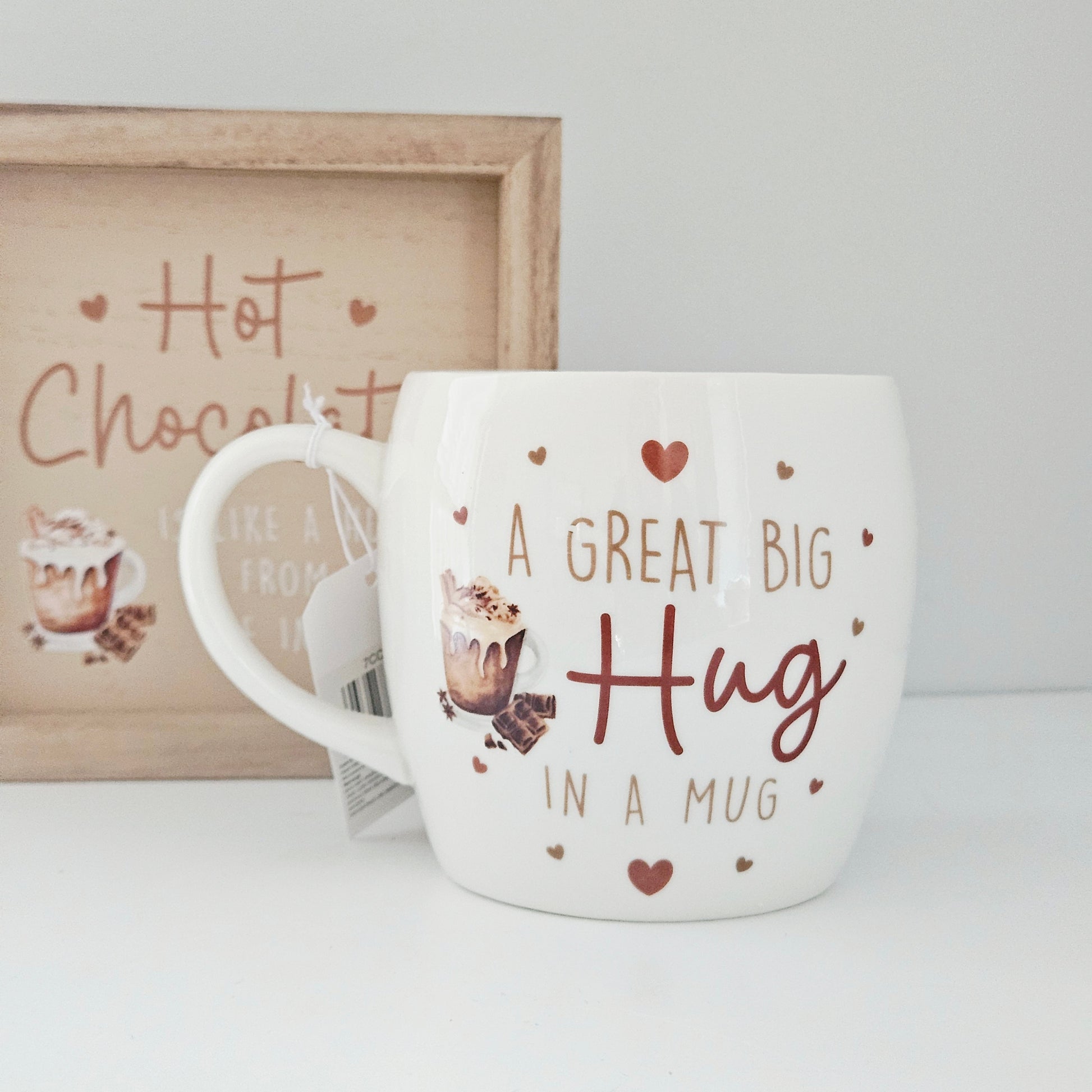 White mug with 'A Great Big Hug in a Mug' text and heart designs, placed on a white surface with a framed picture in the background.