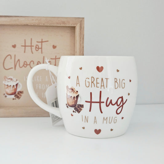 White mug with 'A Great Big Hug in a Mug' text and heart designs, placed on a white surface with a framed picture in the background.