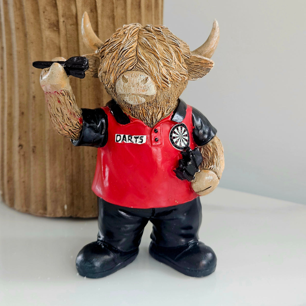A Highland cow figurine wearing a red and black polo shirt with 'DARTS' written on the chest, holding a dart