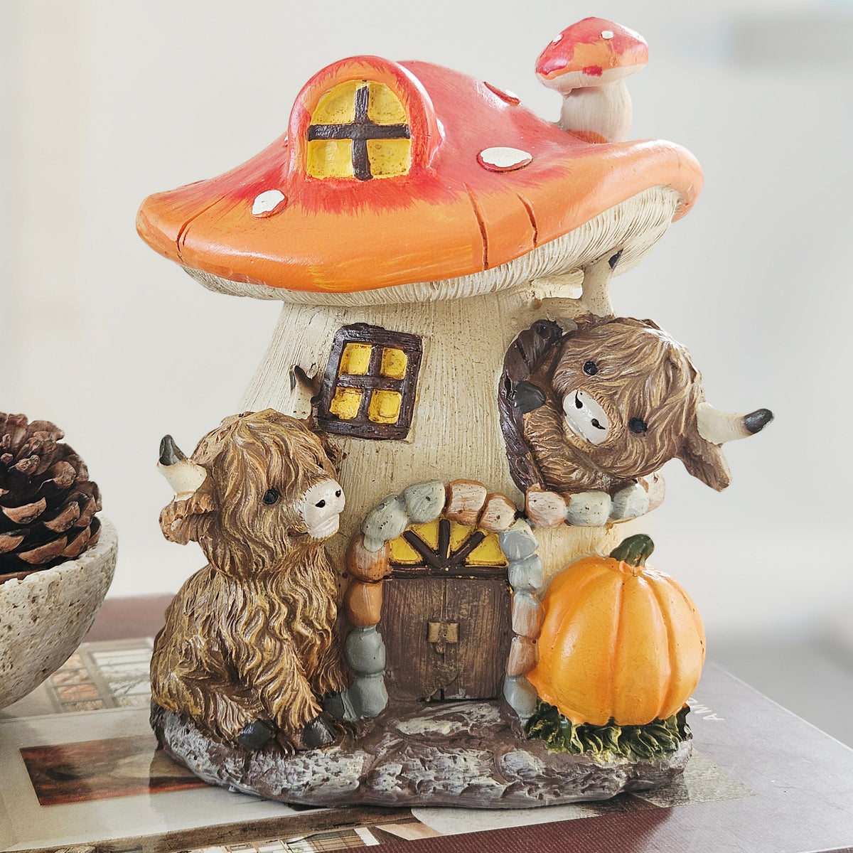 A decorative mushroom-shaped cottage figurine with a red-orange mushroom cap, cream base, small window, door, and two shaggy highland cow characters