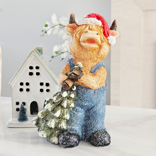 highland cow with red santa hat and blue dungarees on carrying a green christmas tree