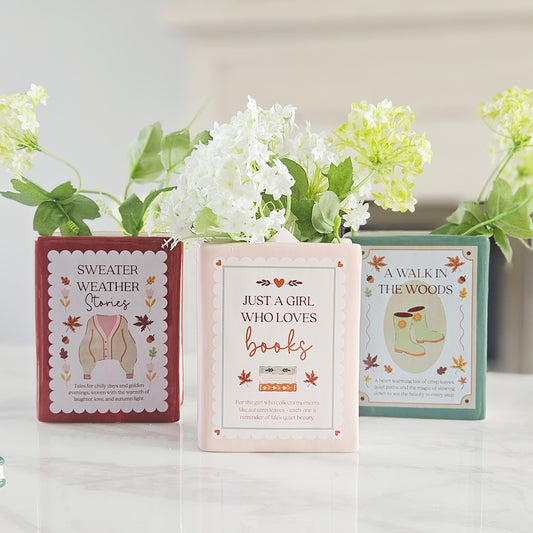 Three decorative book-themed book designed vases. The center box is pale pink and reads 'Just a Girl Who Loves books'; the left box is dusty rose and reads 'Sweater Weather'; the right box is mint green and reads 'A Walk in the Woods'.