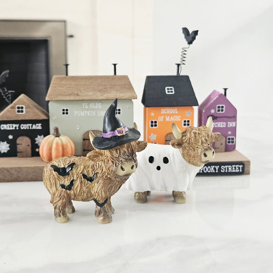 Two highland cow figurines, one wearing a witch hat and the other a ghost costume, with Halloween-themed houses in the background.