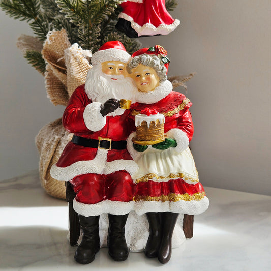 A Christmas figurine depicting Santa Claus and Mrs. Claus sitting together on a bench, holding a small cake.