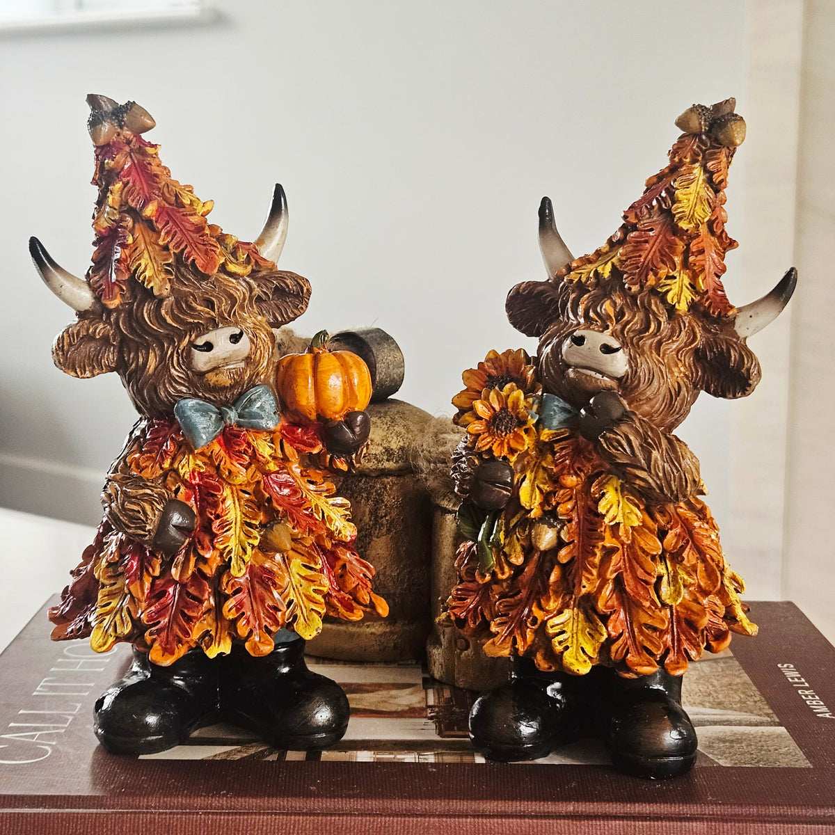Two decorative Highland cow figurines dressed in autumn leaf costumes, holding pumpkin and sunflower