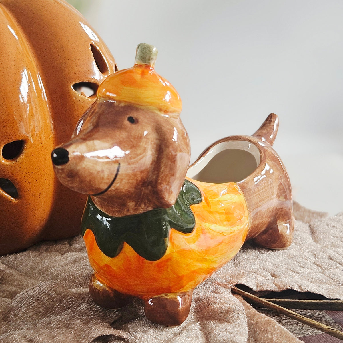 A ceramic dachshund-shaped planter or dish, decorated to look like a dachshund wearing a yellow acorn hat and a green leafy collar over an orange and yellow pumpkin body