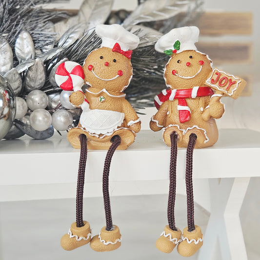 Two gingerbread cookie shelf sitter holiday figurines One wears a white chef's hat and apron holding a red and white striped candy cane, and the other wears a white chef's hat and scarf holding a sign that says 'JOY' in red letters.