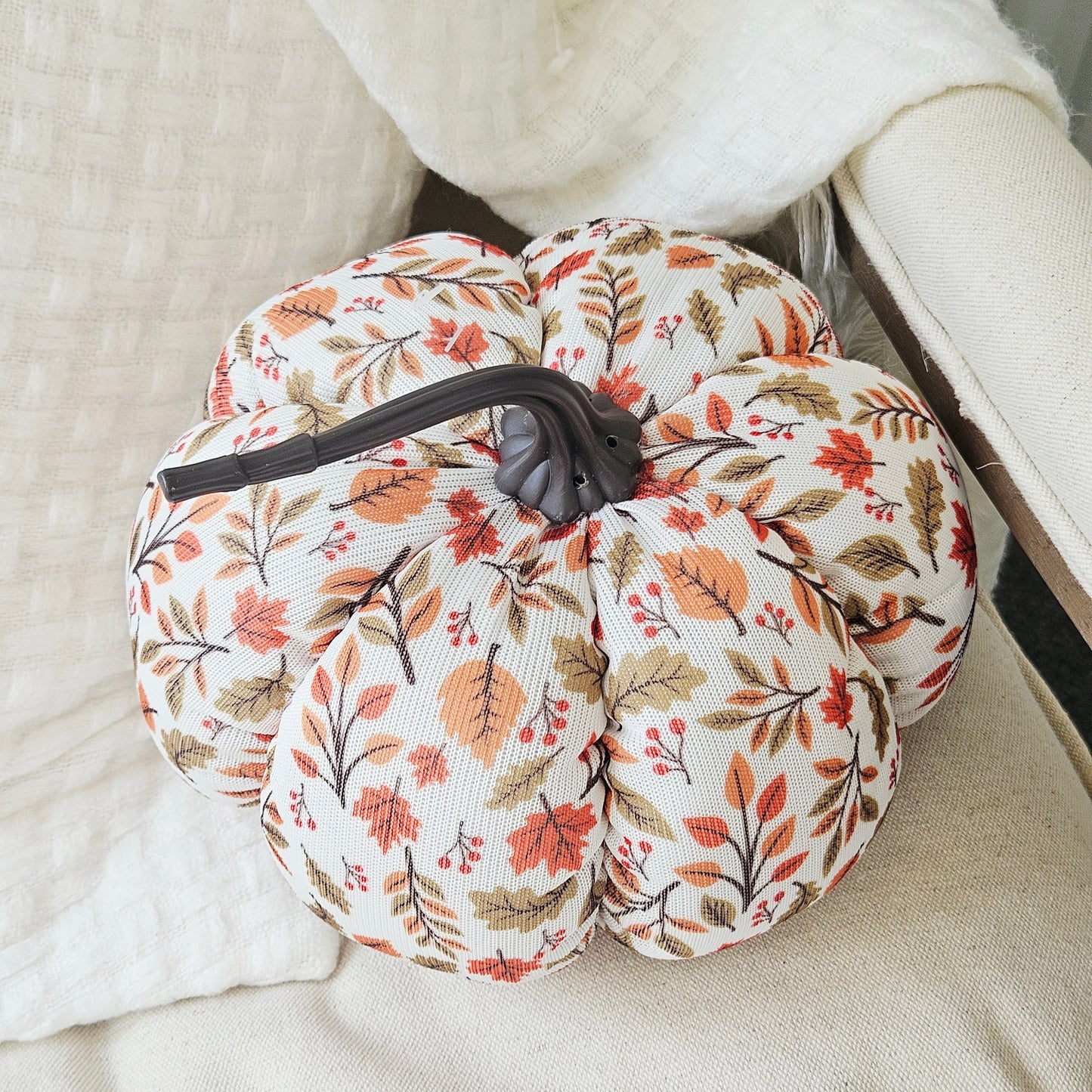 A fabric-covered decorative pumpkin with a fall leaf pattern in orange, brown, and beige on an off-white background, featuring a dark gray stem, styled on a textured cream fabric surface.