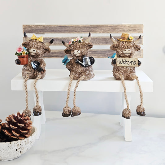 Three decorative Highland cow figurines on a bench flowers, watering can and welcome shelf sitters