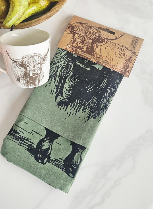 2 Green tea towels with a printed design of a highland cow displayed with a cardboard holder 
