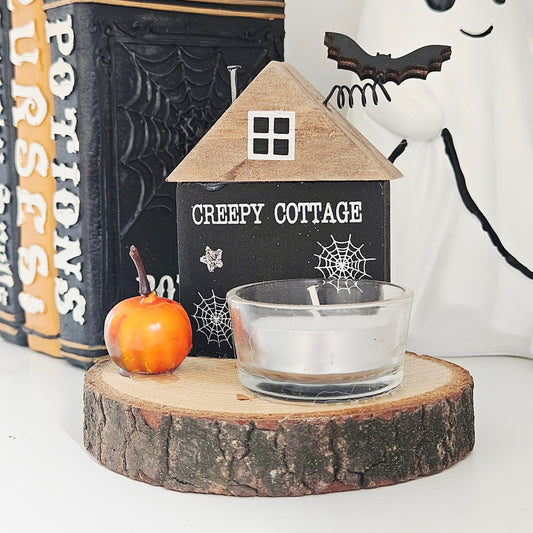 Halloween-themed decor with a 'Creepy Cottage' sign, candle, and pumpkin on a wooden base.
