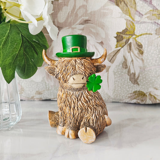 Decorative figurine of a highland cow wearing a green top hat and holding a four-leaf clover, with flowers in mouth