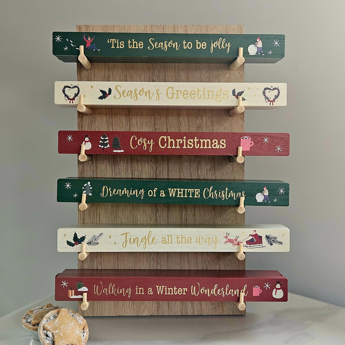 A wooden Christmas sign board with six horizontal planks in dark green and muted red, each with a small wooden hanger holding a matching cream tag. The planks are decorated with festive illustrations and phrases including 'Tis the Season to be jolly', 'Season & Greetings', 'Cosy Christmas', 'Dreaming of a WHITE Christmas', 'Jingle all the way', and 'Walking in a Winter Wonderland'.