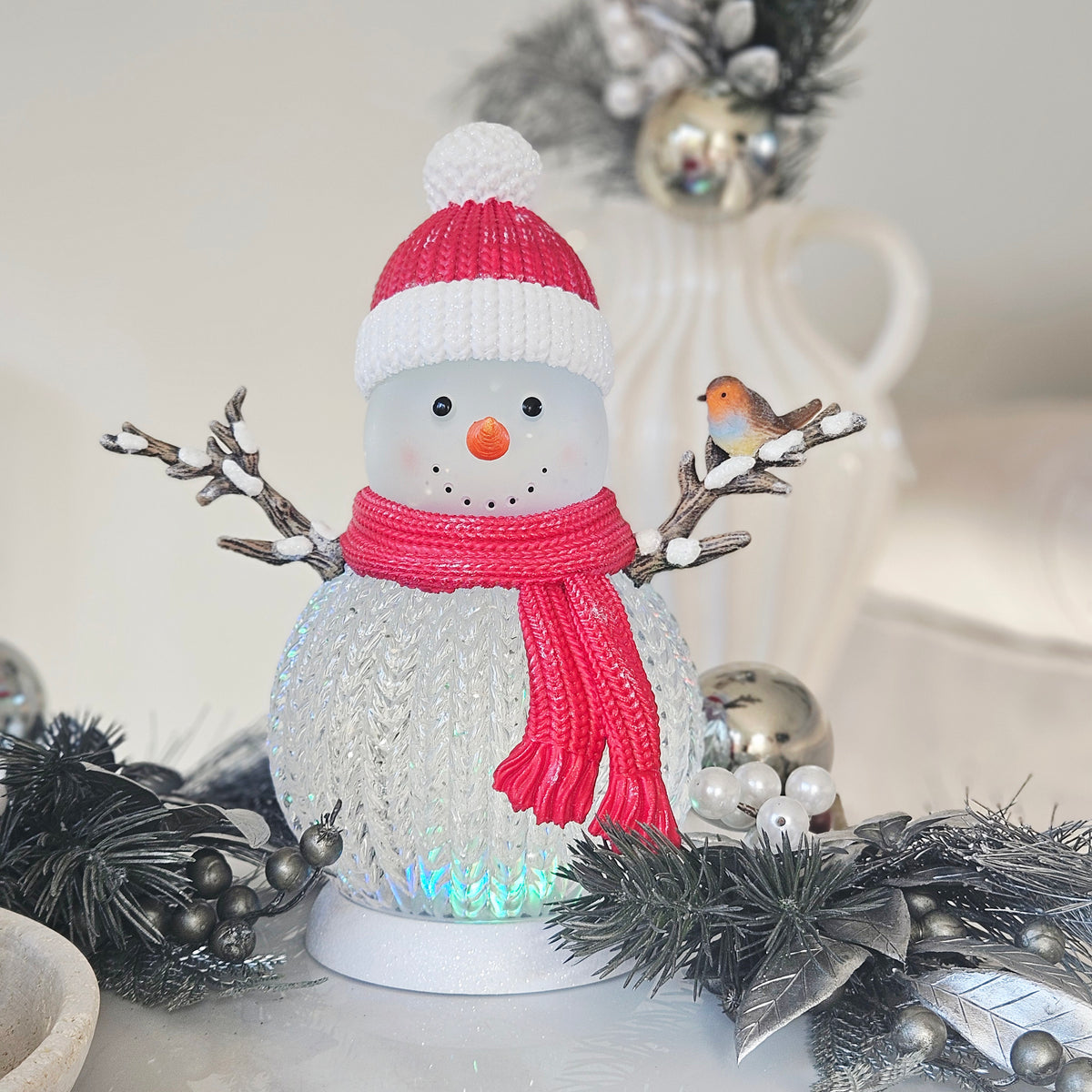 A festive snowman figurine with a red and white knit hat, red scarf, twig arms, and a small robin perched on one twig arms  