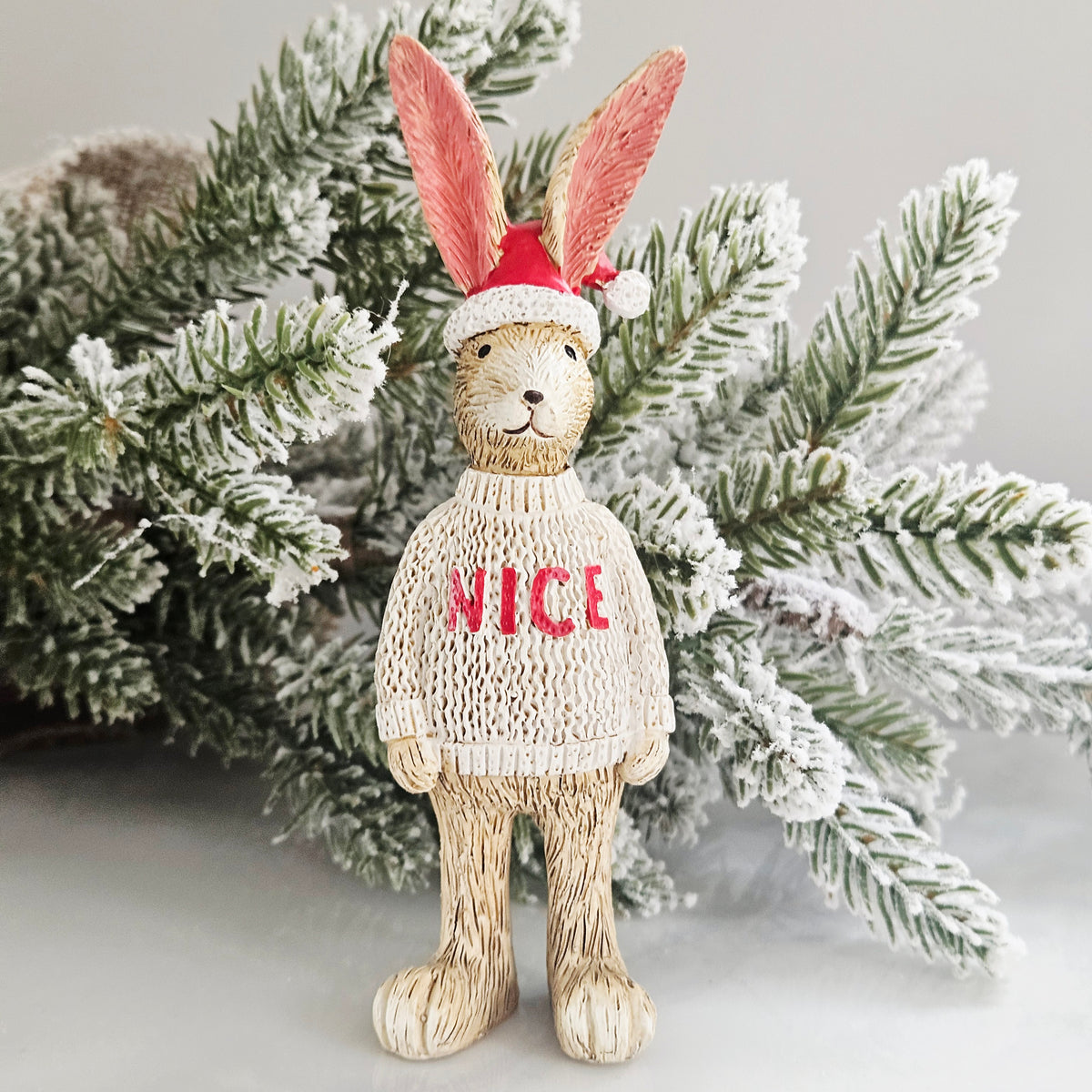 A decorative rabbit figurine wearing a red Santa hat and a cream knit sweater with the word 'NICE' in red,