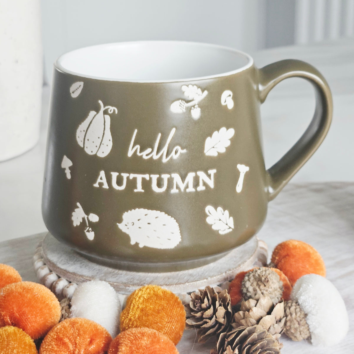 An olive green stoneware mug with a white handle, decorated with white autumn-themed illustrations and the words 'hello AUTUMN'. foreground.