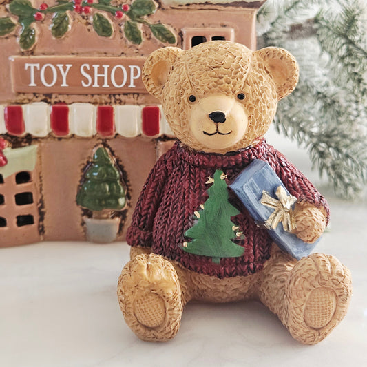 A decorative teddy bear figurine wearing a burgundy knit sweater with a green Christmas tree on the front and holding a blue gift box