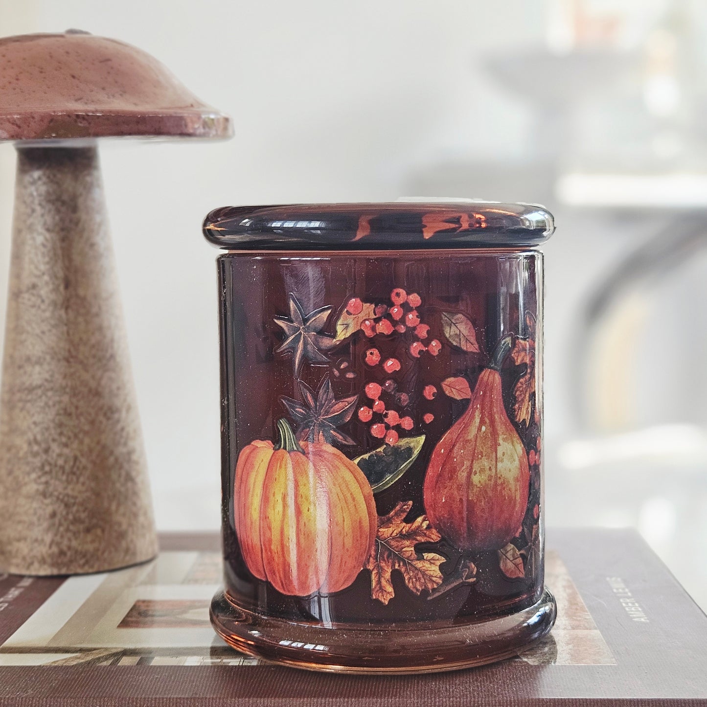 A cylindrical glass jar candle with a lid, decorated with an autumn harvest design featuring pumpkins, pears, berries, and spices on a dark brown background.
