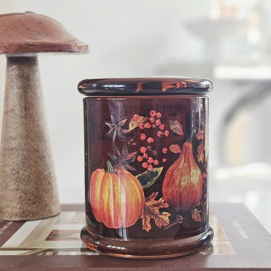 A cylindrical glass jar candle with a lid, decorated with an autumn harvest design featuring pumpkins, pears, berries, and spices on a dark brown background.