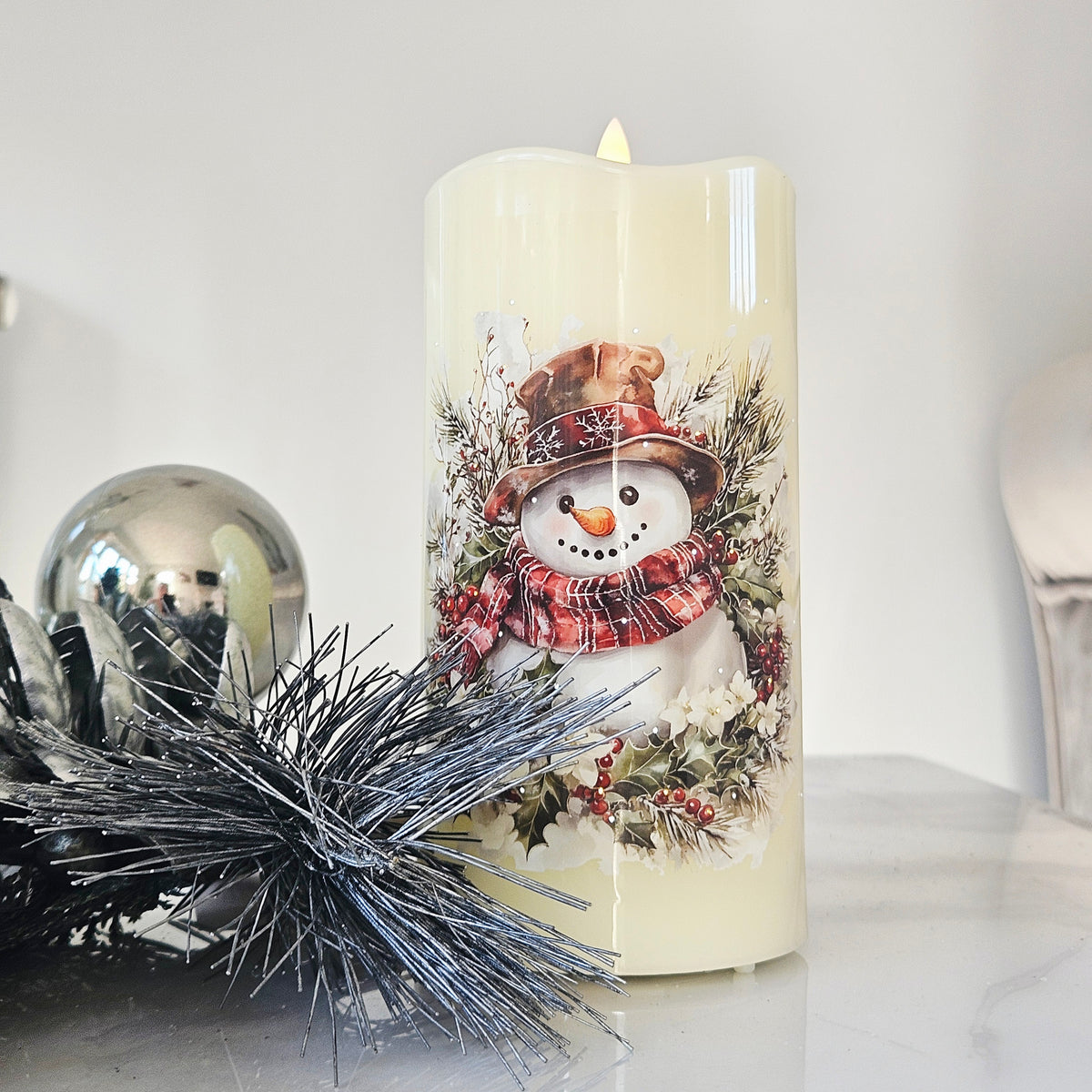 A cream-coloured pillared candle with a printed snowman design wearing a red plaid scarf and hat 15cm