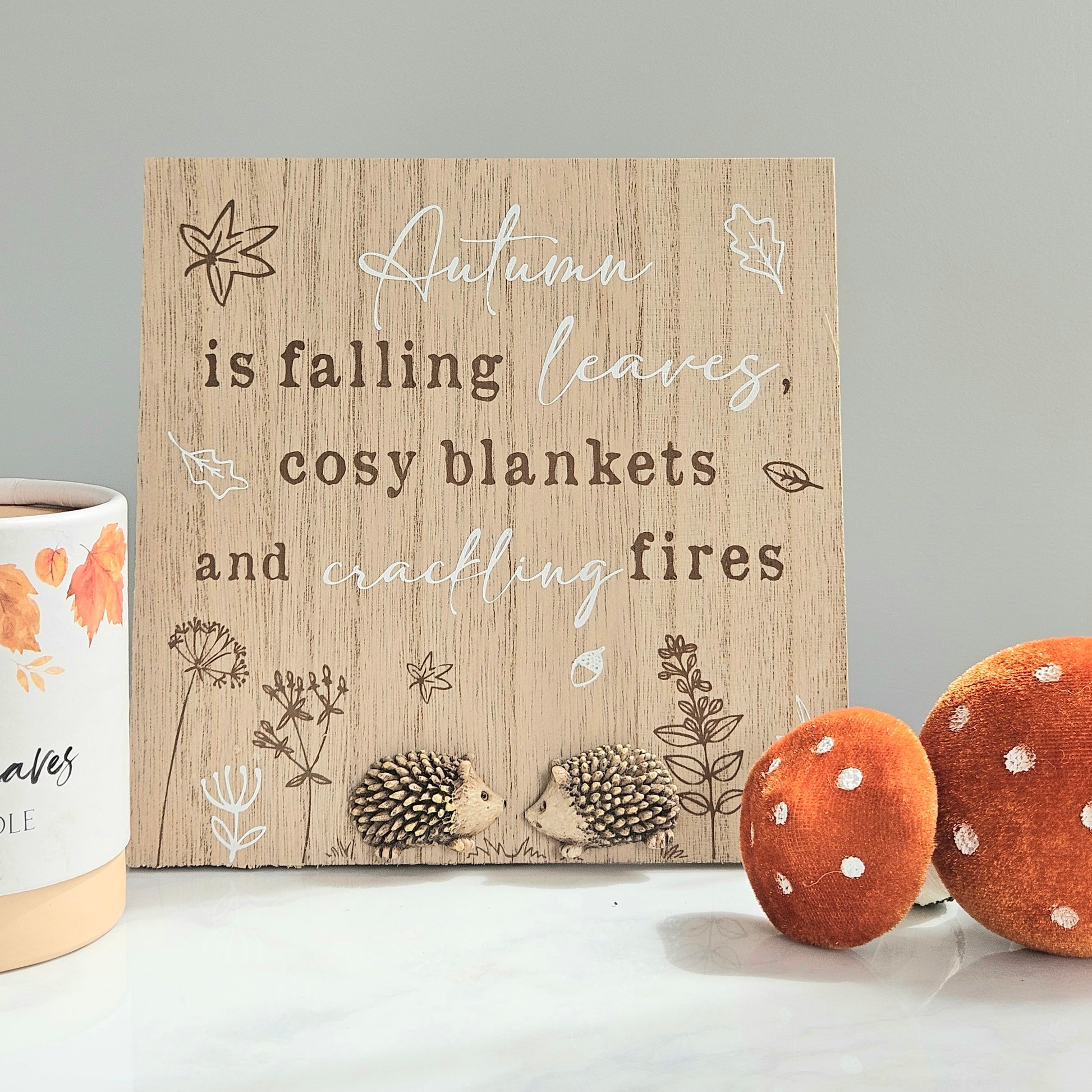 Decorative sign with autumn-themed text and illustrations on a wooden surface with hedgehogs.