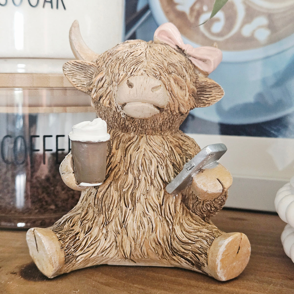 A Highland cow figurine with shaggy tan fur, a pink bow on its head, and a textured mug in one hand and a smartphone in the other,