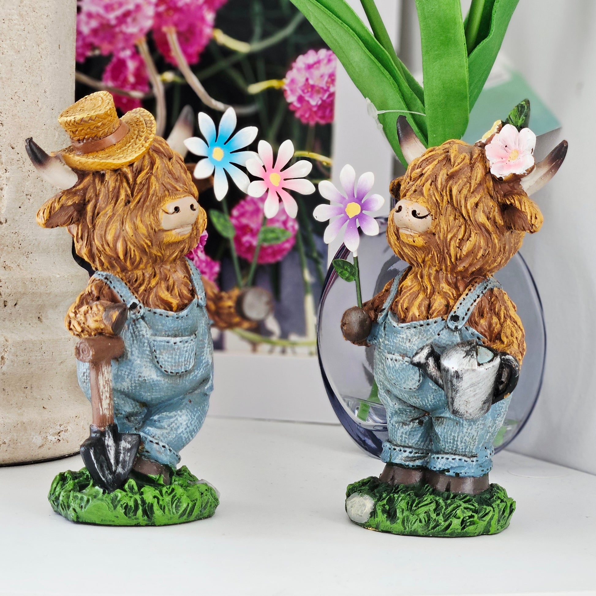 Two decorative figurines of Highland cows with hats and overalls holding flowers