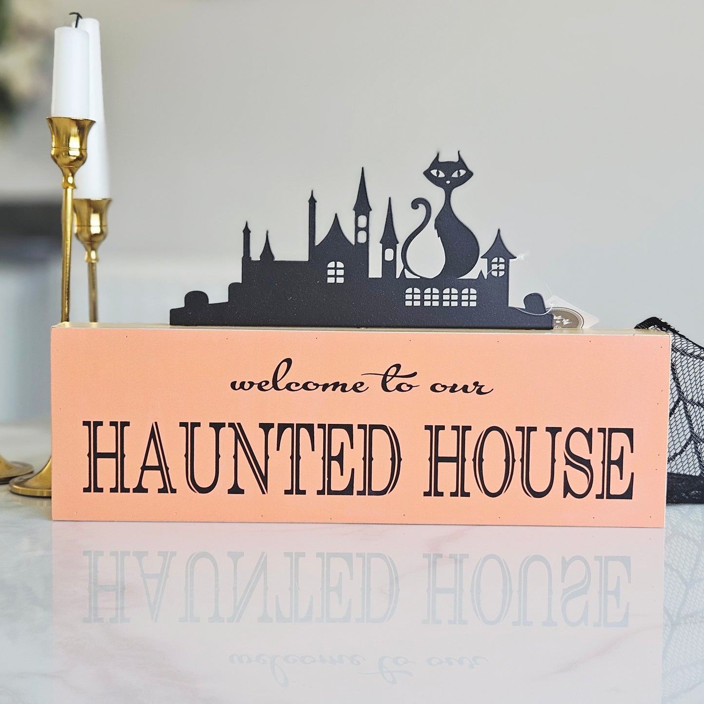 A rectangular orange decorative sign with black text reading 'welcome to our HAUNTED HOUSE' and a black silhouette skyline of a haunted house with a cat on top,