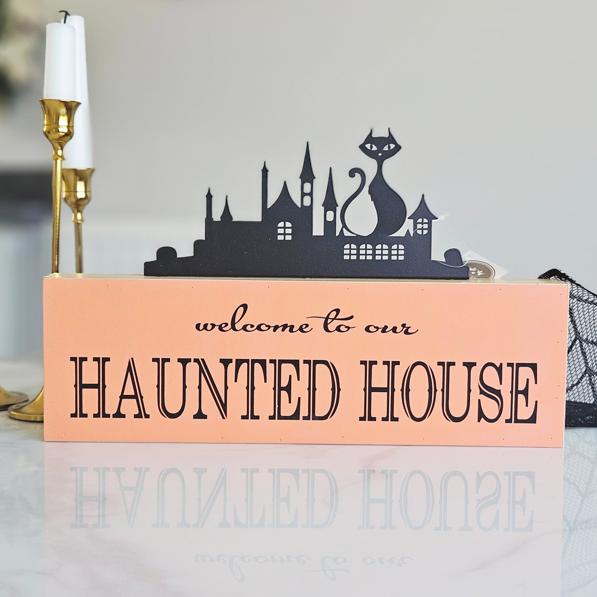 A rectangular orange decorative sign with black text reading 'welcome to our HAUNTED HOUSE' and a black silhouette skyline of a haunted house with a cat on top,
