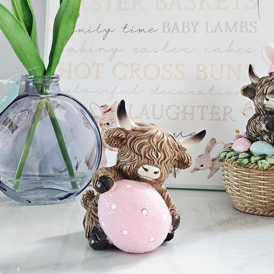 Decorative highland cow figurine with a holding a pink easter egg 
