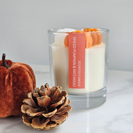 Scented candle labeled 'Spiced Pumpkin & Patchouli' with decorative pumpkins and a pine cone on a light background
