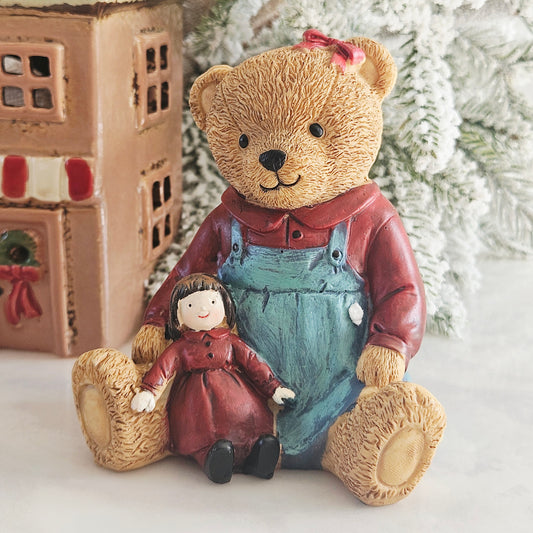 A teddy bear figurine wearing a red shirt and light blue overalls, sitting and holding a small doll with a red dress and brown hair,