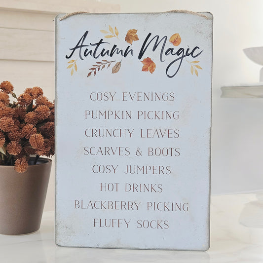A rustic white wooden sign with 'Autumn Magic' written in black script, decorated with small orange and yellow leaves. Below, a list of cozy autumn activities is printed in a muted brown font, including pumpkin picking, scarves and boots, and hot drinks.