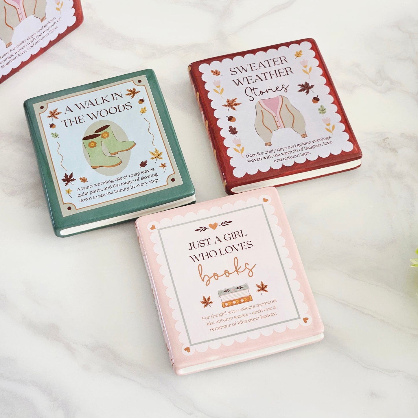 Three hardcover book coasters with autumn-themed covers are arranged on a light marble surface. The covers read "A WALK IN THE WOODS," "SWEATER WEATHER Stories," and "JUST A GIRL WHO LOVES books," with pastel green, rust red, and pale pink designs and fall leaf accents.