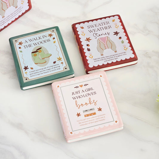 Three hardcover book coasters with autumn-themed covers are arranged on a light marble surface. The covers read "A WALK IN THE WOODS," "SWEATER WEATHER Stories," and "JUST A GIRL WHO LOVES books," with pastel green, rust red, and pale pink designs and fall leaf accents.