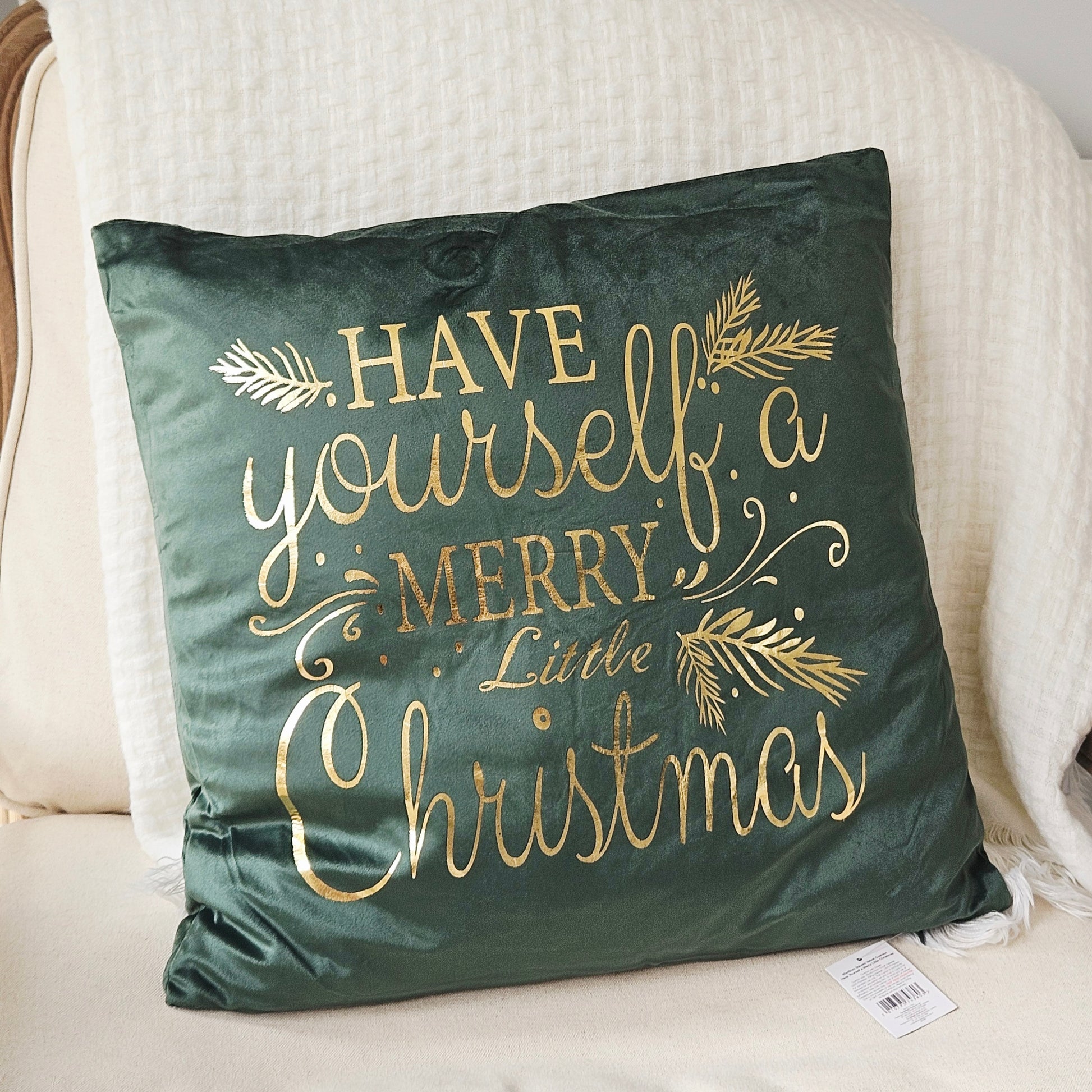 A square dark green velvet throw pillow with gold foil lettering that reads 'HAVE yourself a MERRY Little Christmas' and gold pine branch accents, resting on a cream-colored armchair.