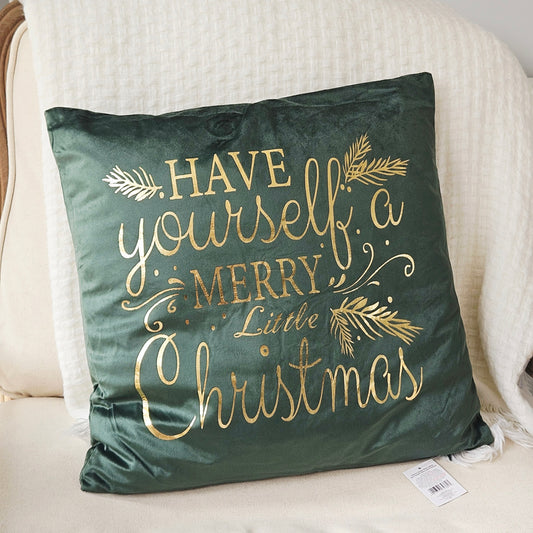 A square dark green velvet throw pillow with gold foil lettering that reads 'HAVE yourself a MERRY Little Christmas' and gold pine branch accents, resting on a cream-colored armchair.