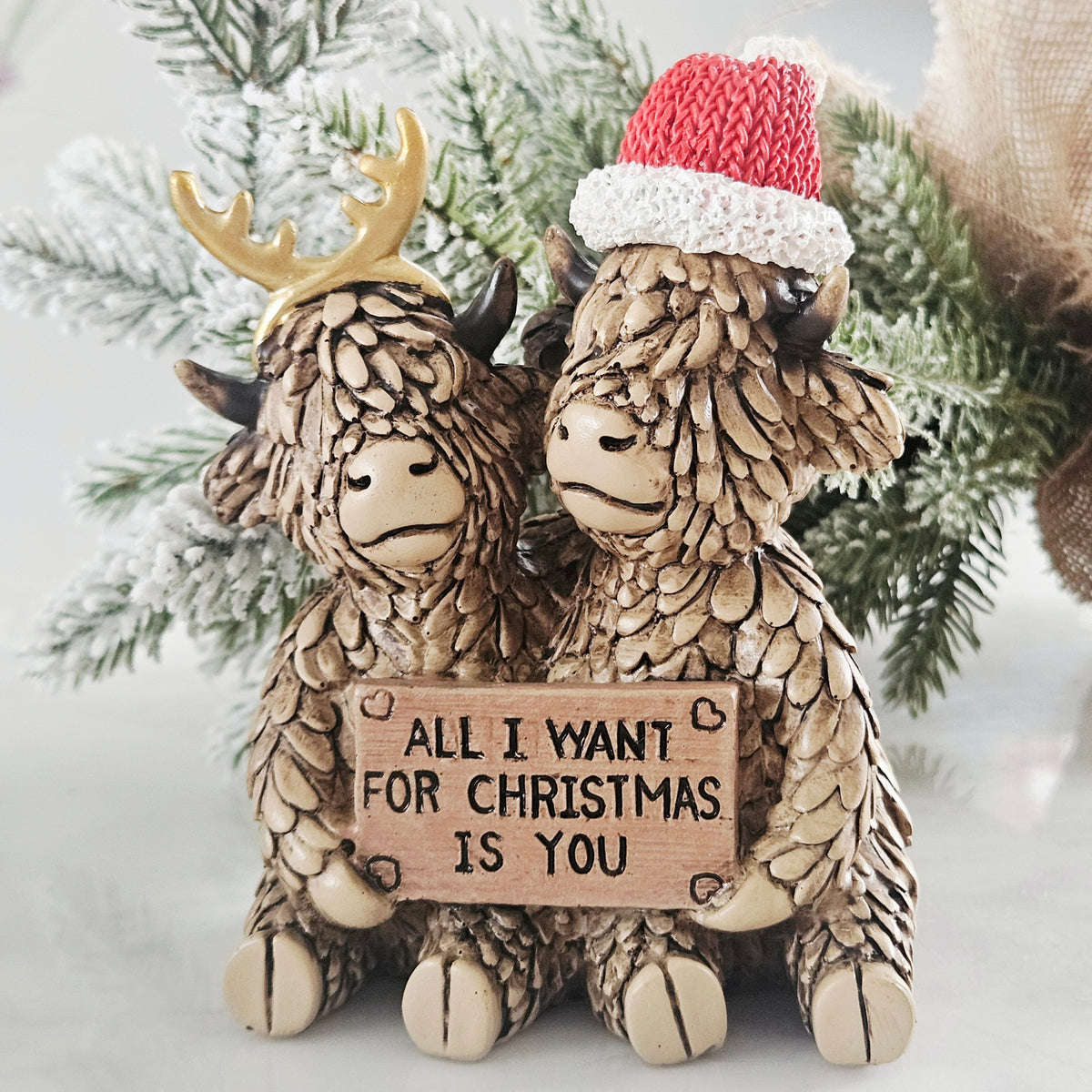 A Christmas-themed decorative figurine of two Highland cows with textured fur. One cow wears a gold reindeer antler and the other wears a red and white knit Santa hat. holding plaque 