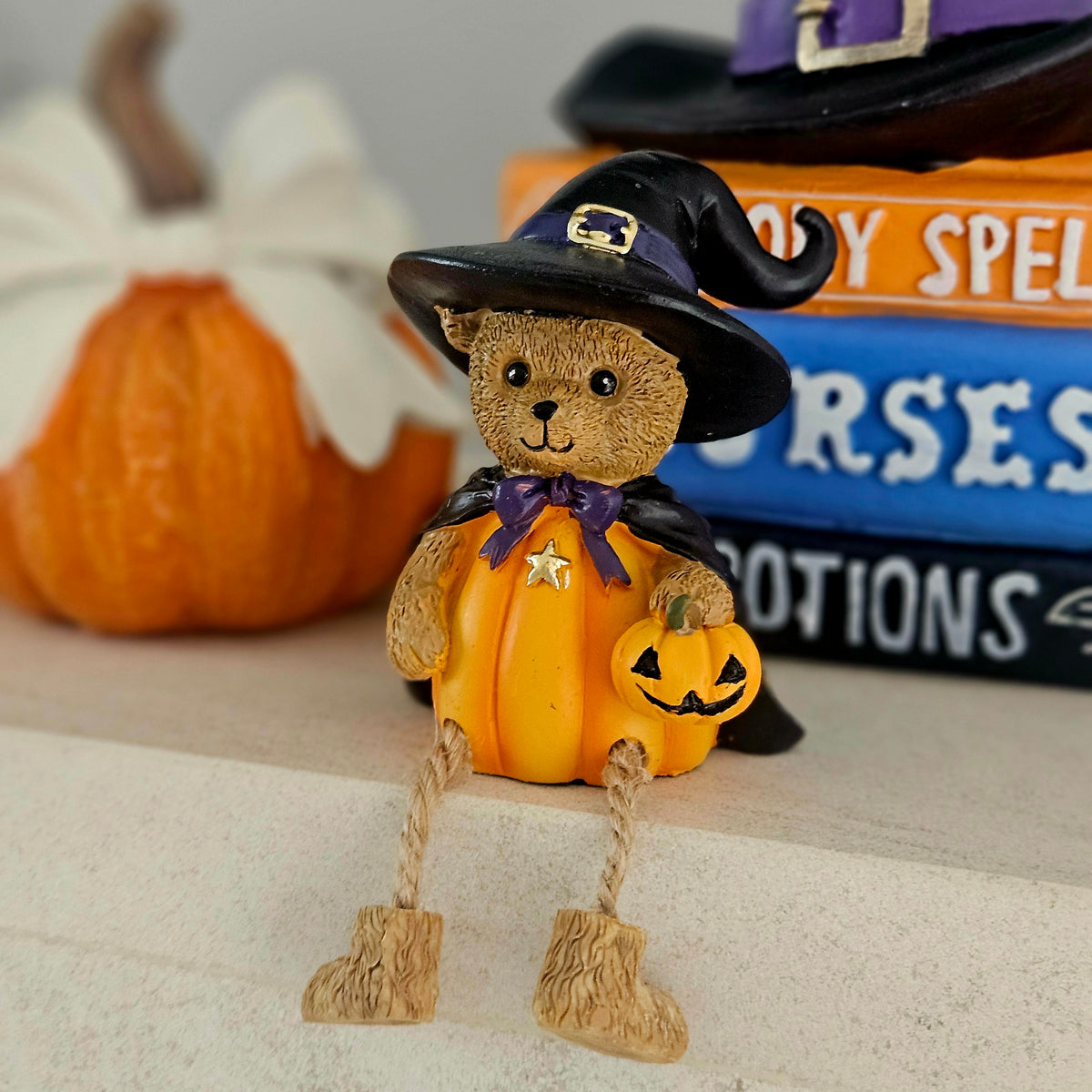 A small Halloween decorative figurine shaped like a teddy bear dressed as a witch, wearing a black hat with a gold buckle, purple cape, and orange pumpkin-shaped outfit, 