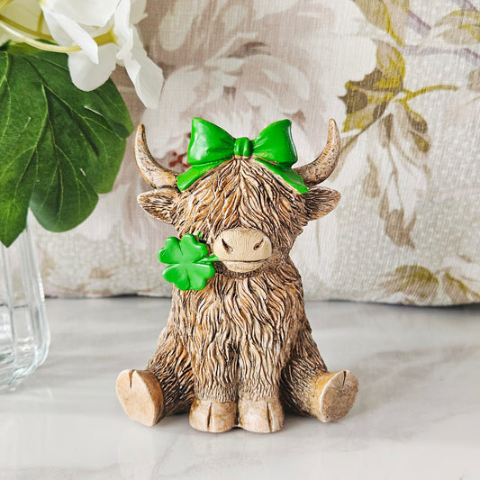 Decorative lucky girl figurine of a Highland cow with a green bow and clover, in her mouth 