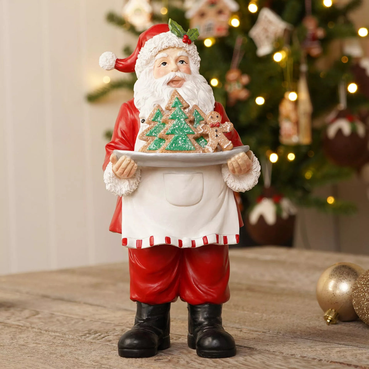 A Santa Claus figurine wearing a red suit and white apron, holding a tray with decorated Christmas trees and gingerbread cookies